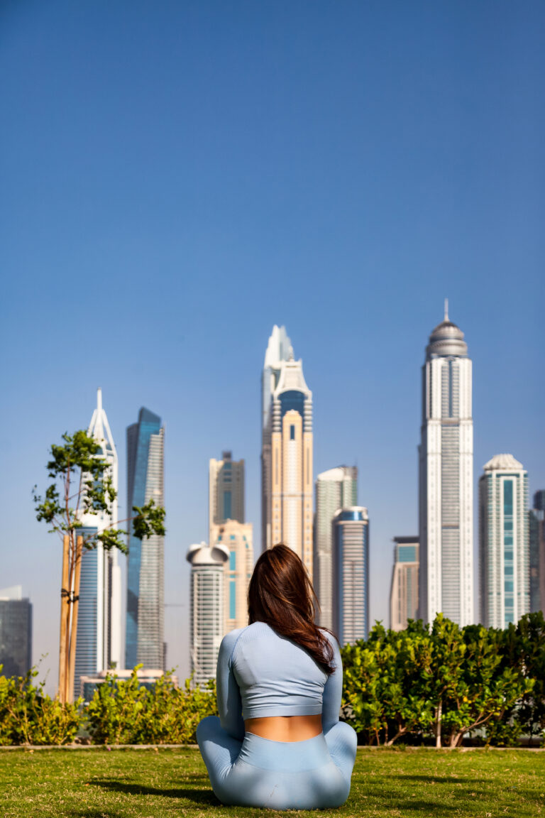 Rear view of sporty adult lady sitting lotus yoga pose on green lawn at Dubai skyscrapers